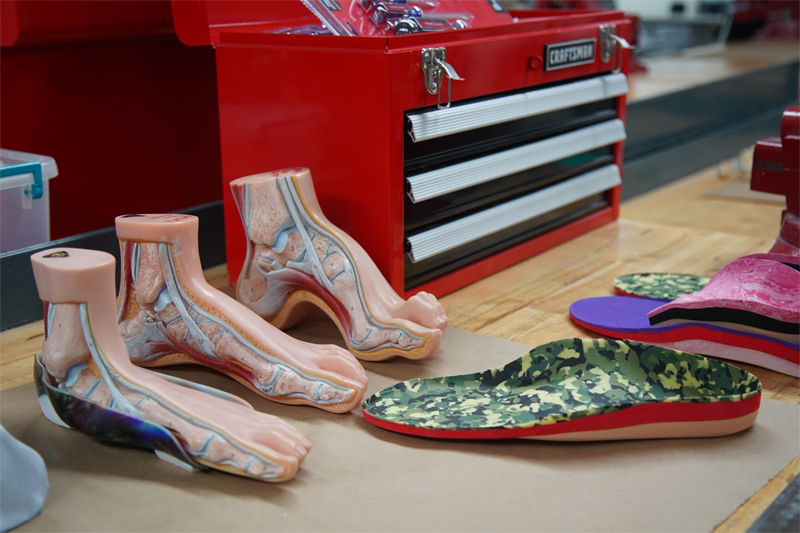 Three anatomy models of the feet are displayed on a workbench with insoles and a toolbox nearby.